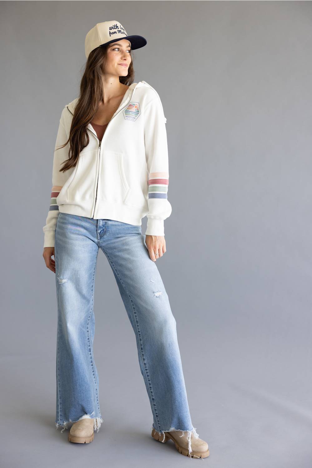 A woman modeling a white zip-up hoodie with colorful stripes on the sleeves, light-wash wide-leg jeans, and a baseball cap against a solid gray studio background.