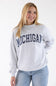A smiling blonde woman wearing a light heather gray crewneck sweatshirt with 'MICHIGAN' printed in navy blue arched letters across the chest, paired with light blue jeans, against a plain white studio background.