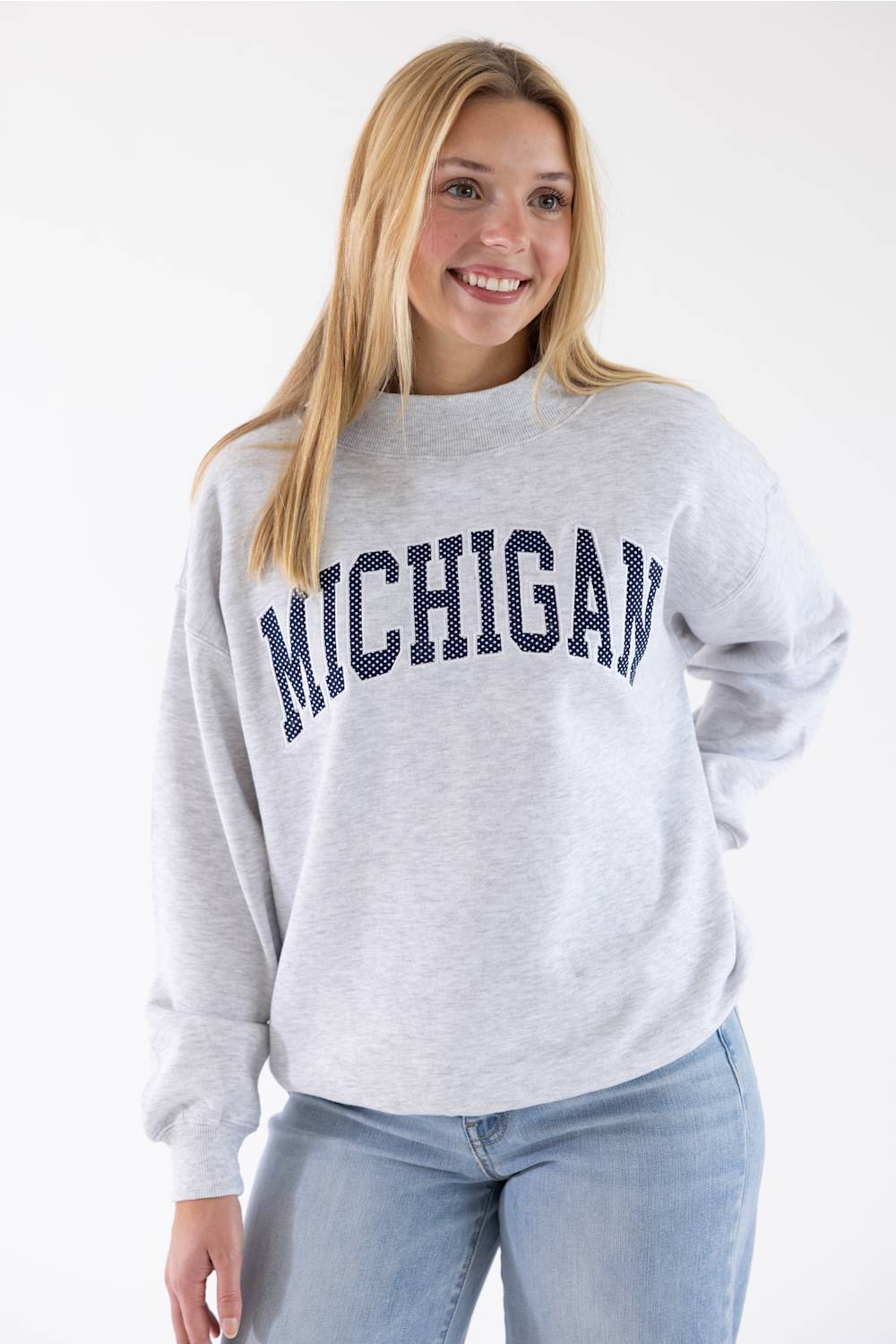 A smiling blonde woman wearing a light heather gray crewneck sweatshirt with 'MICHIGAN' printed in navy blue arched letters across the chest, paired with light blue jeans, against a plain white studio background.