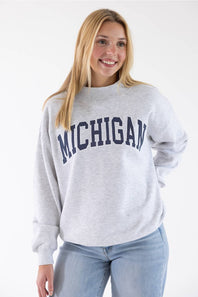 A smiling blonde woman wearing a light heather gray crewneck sweatshirt with 'MICHIGAN' printed in navy blue arched letters across the chest, paired with light blue jeans, against a plain white studio background.