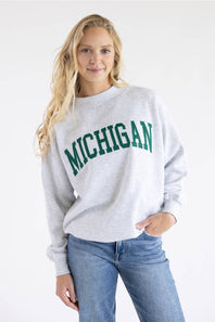 A woman wearing a light gray heather crewneck sweatshirt with 'MICHIGAN' printed in green across the chest, paired with blue jeans, against a plain white background.