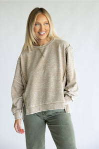 A smiling blonde woman wearing a beige acid-wash crewneck sweatshirt and olive green pants, standing against a light gray studio background.