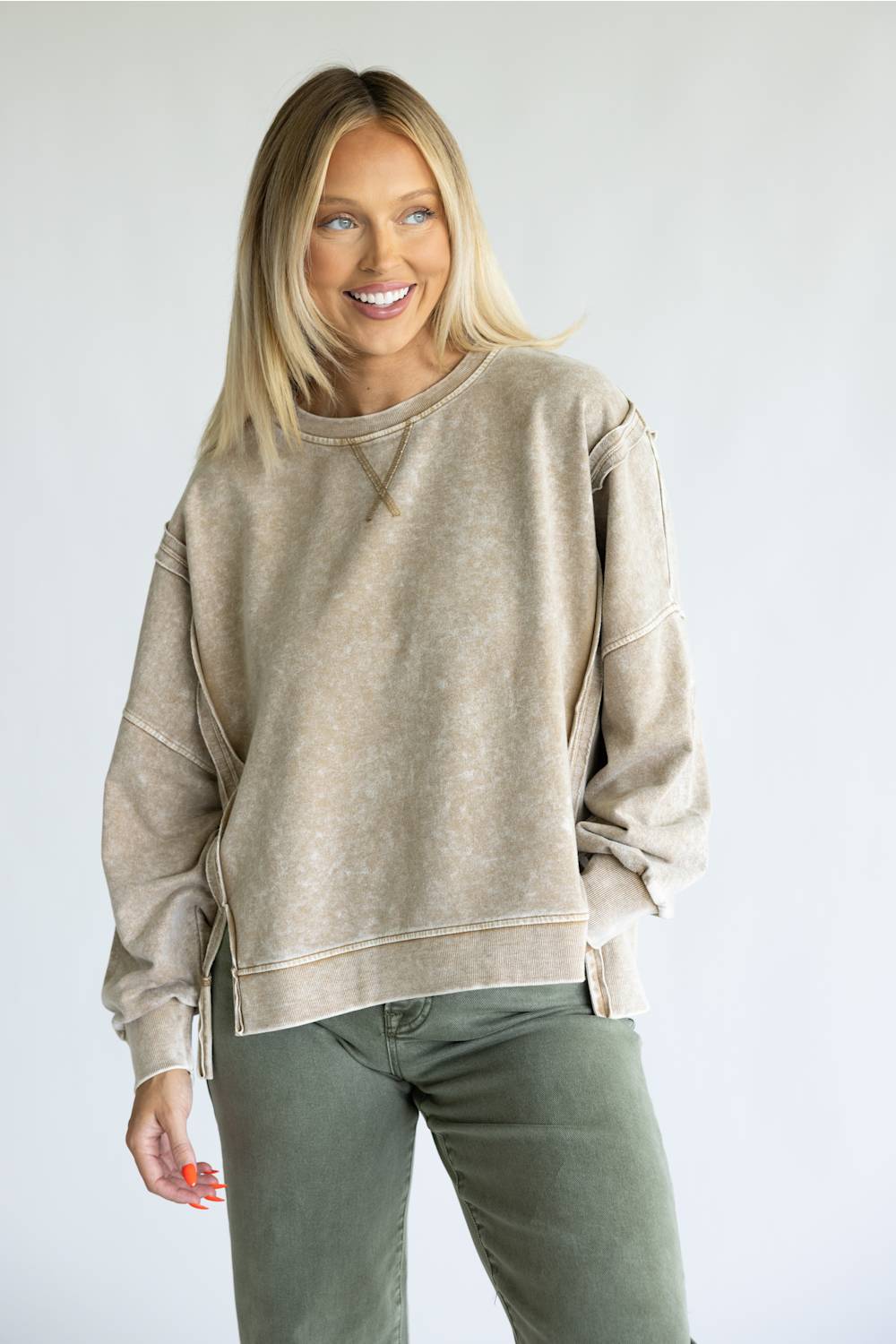 A smiling blonde woman wearing a beige acid-wash crewneck sweatshirt and olive green pants, standing against a light gray studio background.