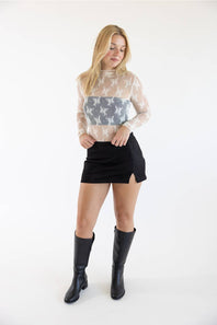 A woman modeling a sheer white long-sleeve top with a gray star pattern across the chest, paired with a black mini skirt and black knee-high boots, against a plain white studio background.