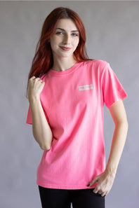 A woman with red hair wearing a bright pink short-sleeve t-shirt with a small rectangular logo patch on the left chest, paired with black pants, against a plain gray studio background.