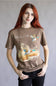 A woman with red hair wearing a brown graphic t-shirt featuring ducks and reeds, paired with black pants, standing against a neutral gray background.