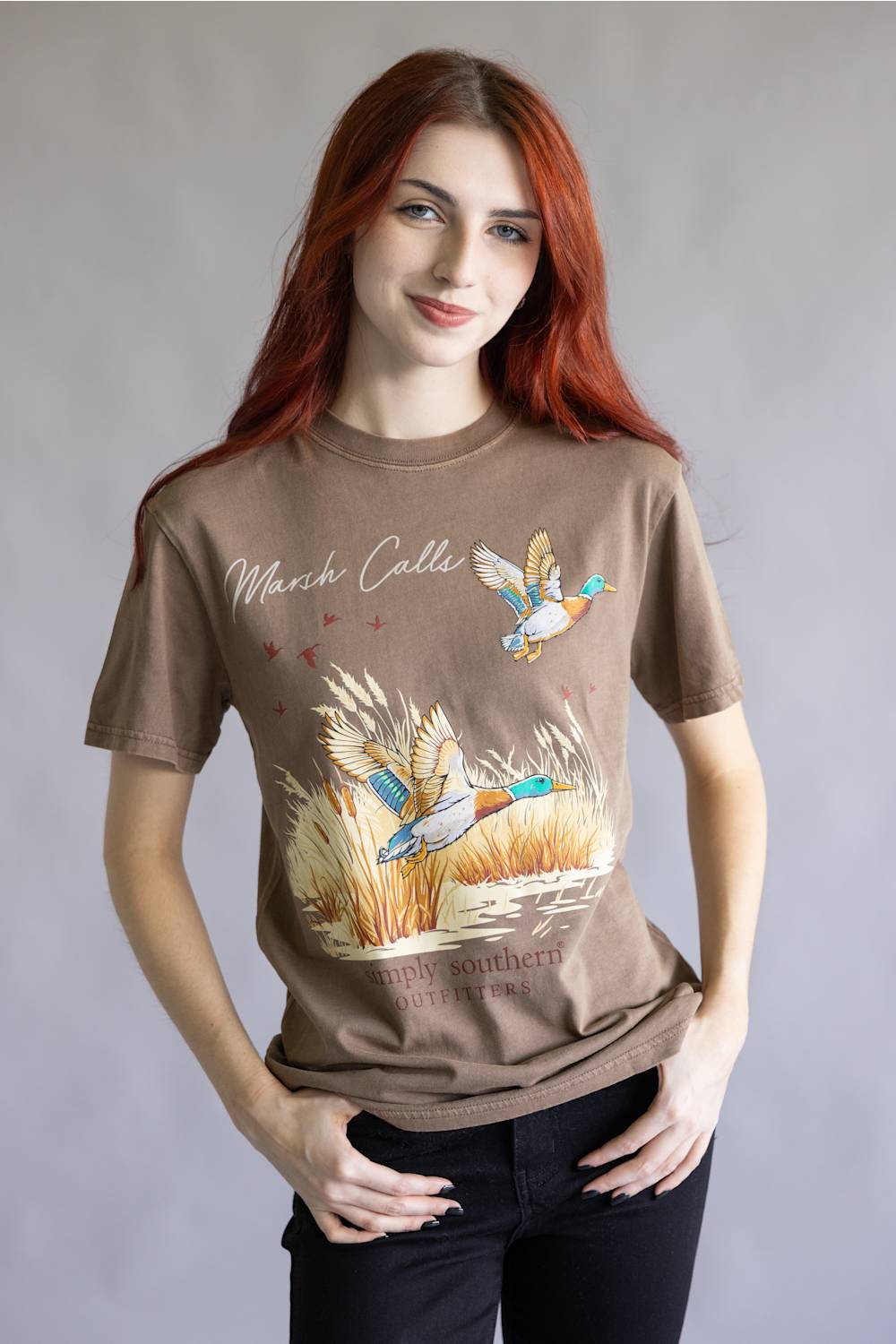 A woman with red hair wearing a brown graphic t-shirt featuring ducks and reeds, paired with black pants, standing against a neutral gray background.