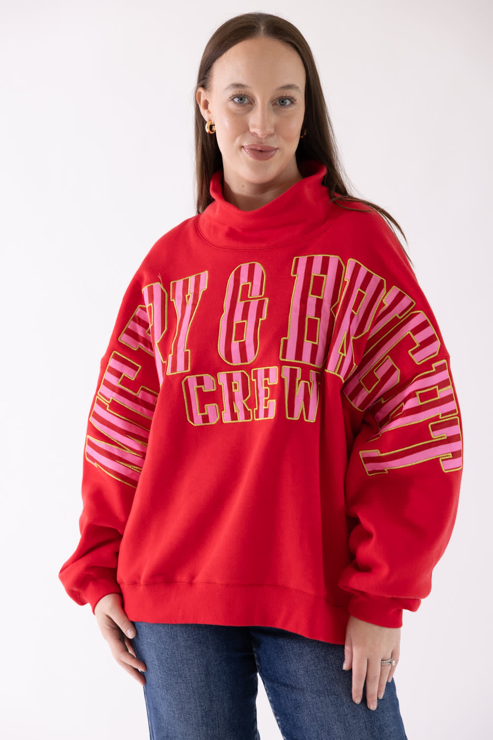 A woman wearing a bright red sweatshirt with mERRY & bRIGHT text in pink and gold on the front, paired with blue jeans, against a plain white background.