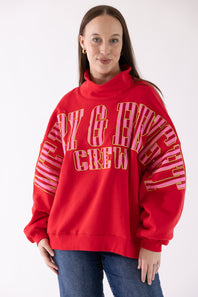 A woman wearing a bright red sweatshirt with mERRY & bRIGHT text in pink and gold on the front, paired with blue jeans, against a plain white background.