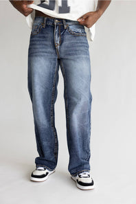 A person wearing a pair of relaxed-fit medium-wash blue denim jeans with whiskering and faded details, styled with a white t-shirt and black and white sneakers against a light gray studio background.