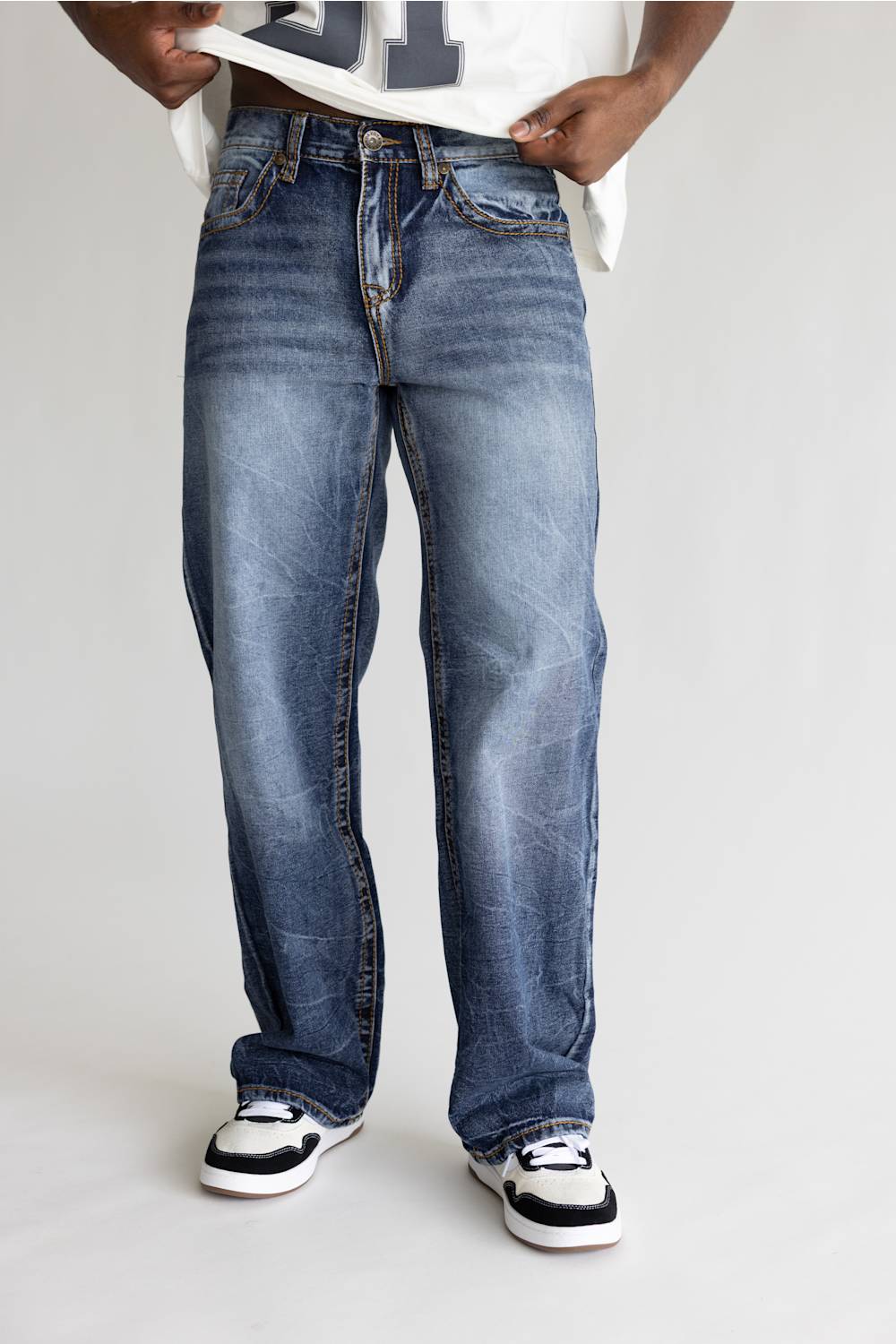 A person wearing a pair of relaxed-fit medium-wash blue denim jeans with whiskering and faded details, styled with a white t-shirt and black and white sneakers against a light gray studio background.