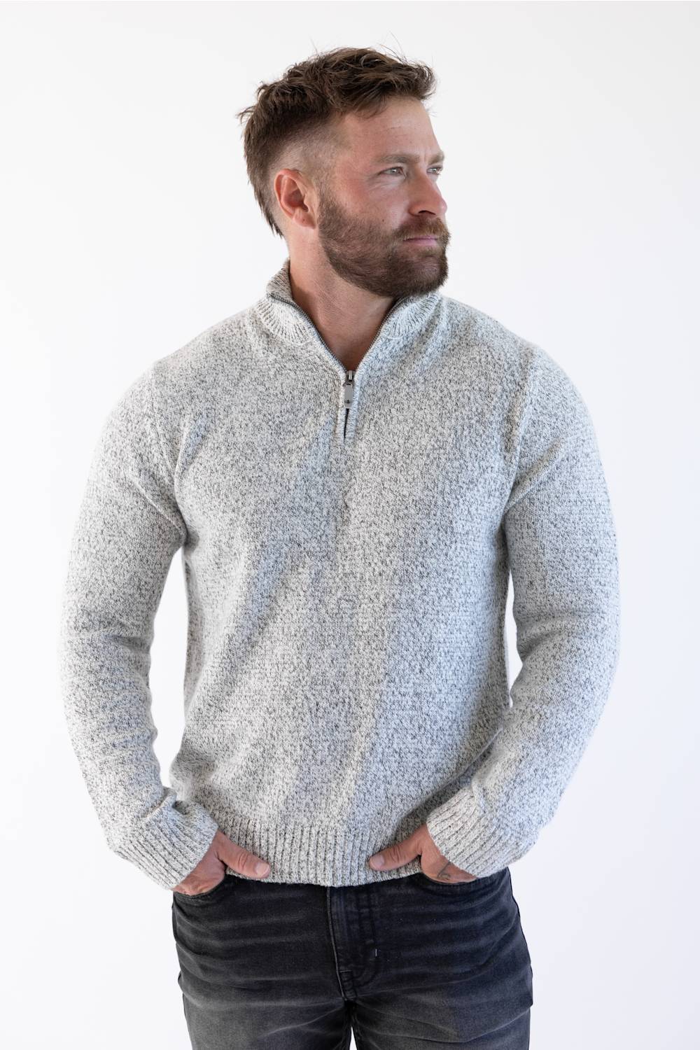 A man wearing a light gray marled quarter-zip knit sweater and dark jeans, standing against a plain white studio background.