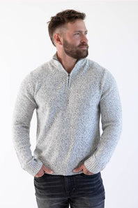 A man wearing a light gray marled quarter-zip knit sweater and dark jeans, standing against a plain white studio background.