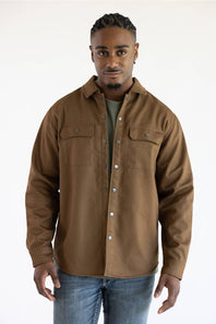 A man wearing a brown button-up shirt jacket over a dark green t-shirt and blue jeans, standing against a plain white studio background.