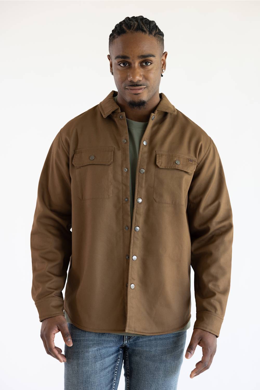A man wearing a brown button-up shirt jacket over a dark green t-shirt and blue jeans, standing against a plain white studio background.