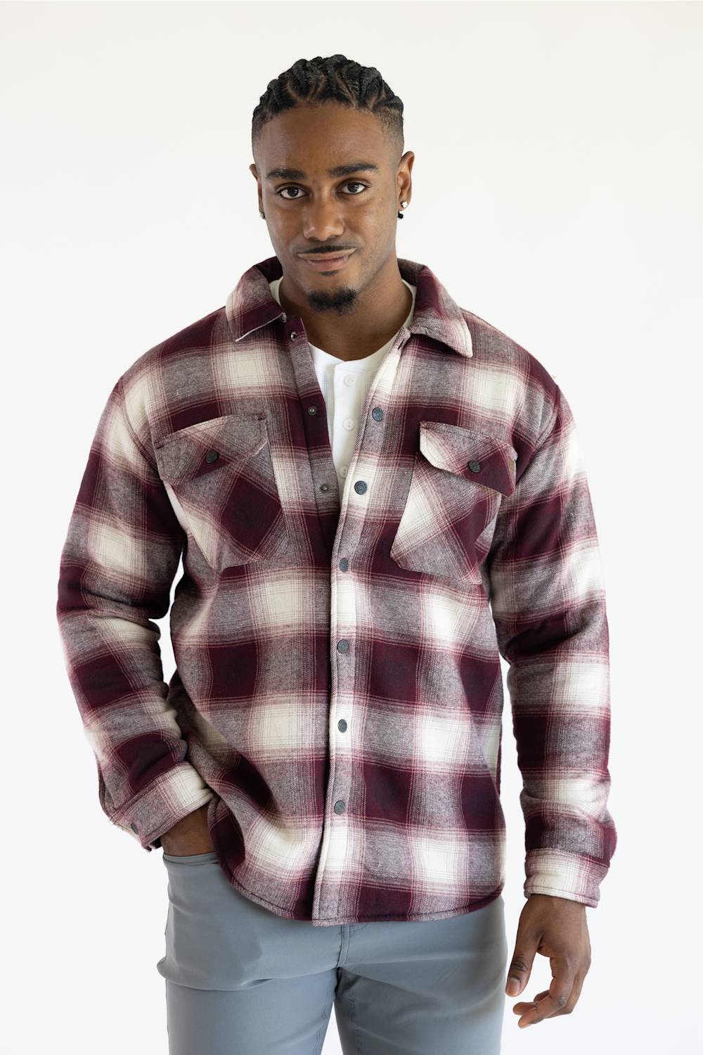 A man wearing a long-sleeve button-up plaid flannel shirt in maroon and white, layered over a white t-shirt and paired with light gray pants, against a plain white background.