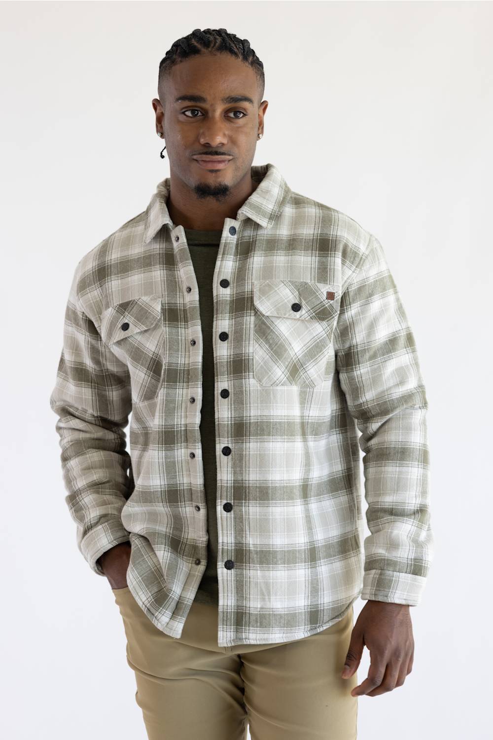 A man wearing a long-sleeve button-up plaid overshirt in beige and olive green, paired with khaki pants, standing against a plain white studio background.