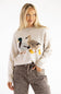 A woman wearing a cream-colored knit sweater with a large embroidered duck family motif on the front, paired with brown leopard-print pants, against a plain white studio background.