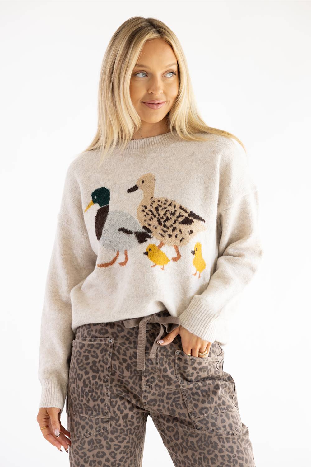 A woman wearing a cream-colored knit sweater with a large embroidered duck family motif on the front, paired with brown leopard-print pants, against a plain white studio background.