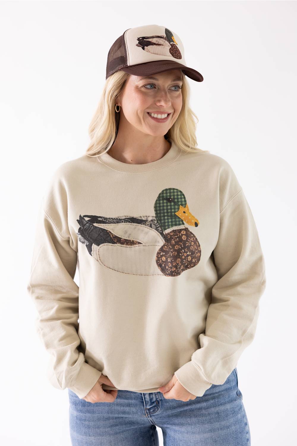Model wearing the Mallard Duck Sweatshirt for Women in Cream and a brown and cream trucker hat with a mallard duck patch, standing against a neutral background.