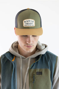 A man wearing a green and tan trucker hat with a 'Marsh Wear' patch on the front, layered over a gray hoodie and a blue-green vest, against a plain white background.