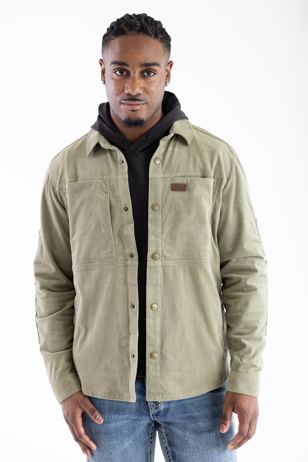 Front studio portrait of a model wearing the Sage Brush green diamond check corduroy Marsh Wear Delano Cord Shacket, featuring an embossed leather brand patch on the left chest and antique brass snap closures, layered over a dark hoodie and styled with blue jeans.