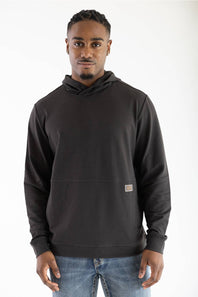 Front-facing studio shot of a model wearing the dark raven black Marsh Wear Fireside Hoodie with the hood down, featuring a crossover neckline, front pouch pocket, and small woven logo patch, styled with light-wash blue jeans.