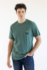 A man wearing a muted green pocket t-shirt with a black graphic on the chest pocket, paired with blue jeans, standing against a plain white background.