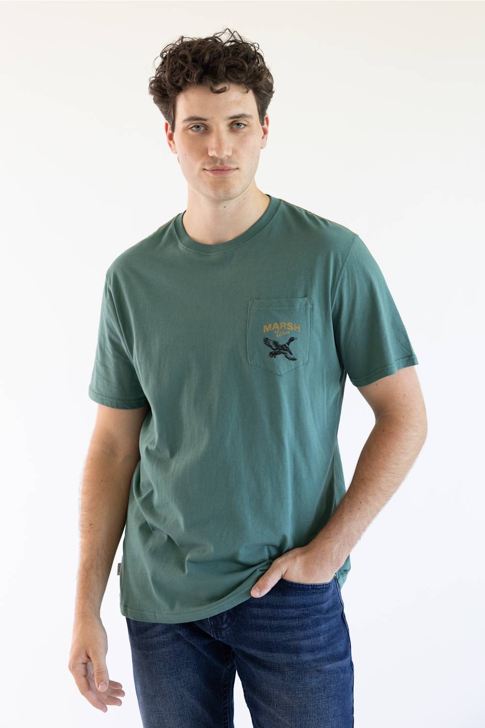 A man wearing a muted green pocket t-shirt with a black graphic on the chest pocket, paired with blue jeans, standing against a plain white background.