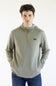 A man wearing a Marshwear hooded quarter-zip pullover in a textured sage green fabric with a small orange zipper and a small Marshwear  logo patch on the chest, paired with blue jeans against a plain white background.
