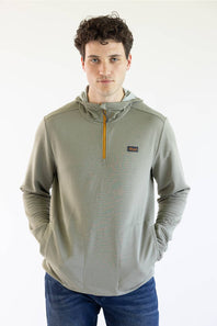 A man wearing a Marshwear hooded quarter-zip pullover in a textured sage green fabric with a small orange zipper and a small Marshwear  logo patch on the chest, paired with blue jeans against a plain white background.
