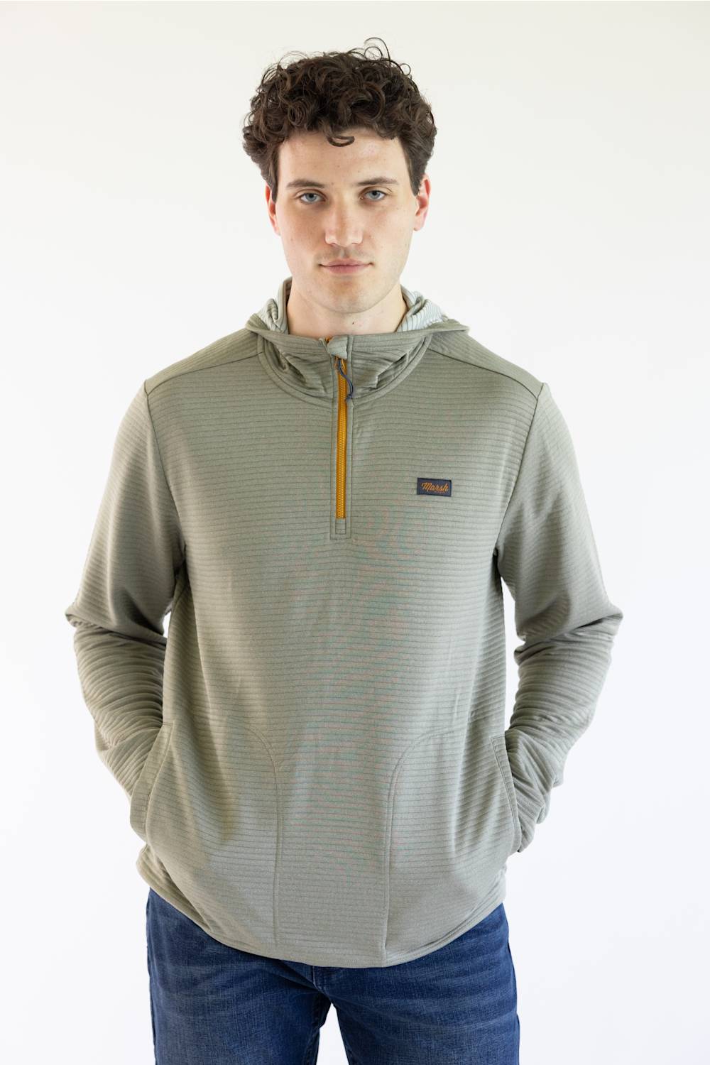 A man wearing a Marshwear hooded quarter-zip pullover in a textured sage green fabric with a small orange zipper and a small Marshwear  logo patch on the chest, paired with blue jeans against a plain white background.