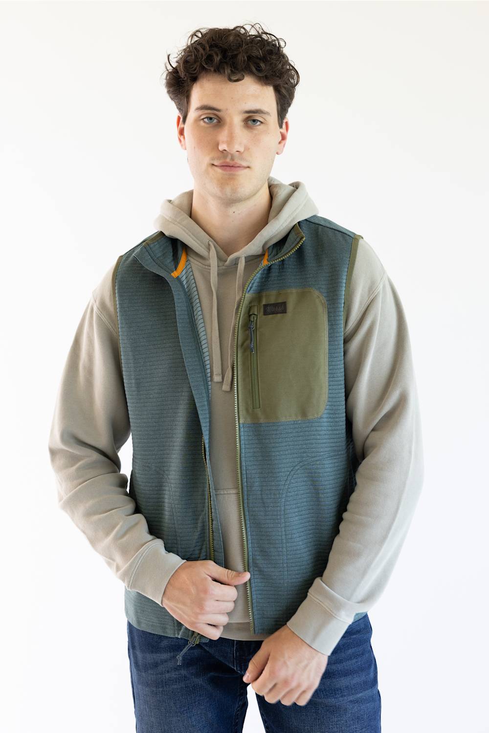 A man wearing a sleeveless zip-front vest over a light gray hoodie and blue jeans, standing against a plain white studio background.