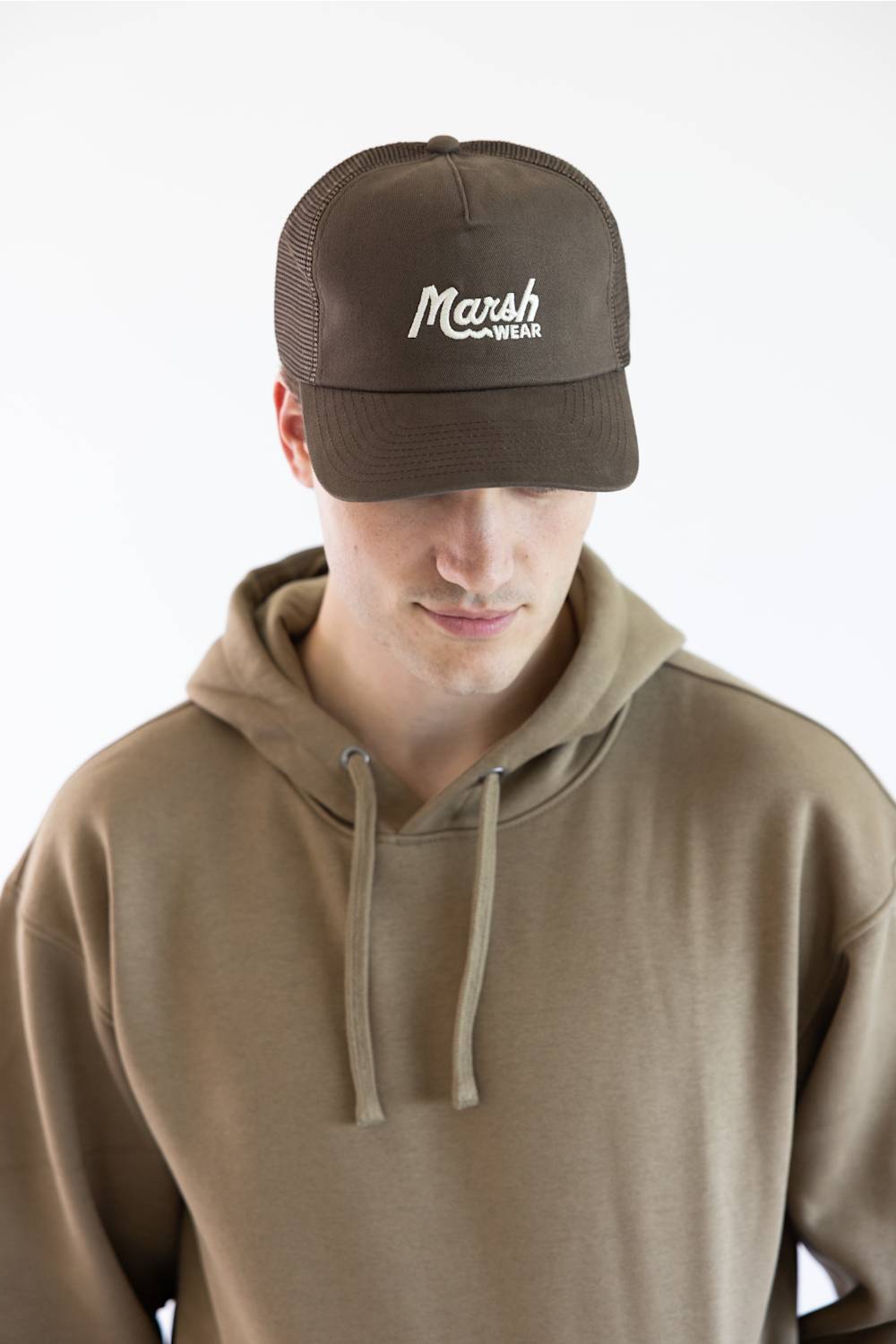 A man wearing a brown trucker hat with 'Marsh Wear' embroidered in white on the front, paired with a brown hoodie, against a plain white background.