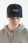 A man wearing a dark navy blue trucker hat with yellow embroidered 'Marsh Wear' text, paired with a light beige hoodie, against a plain white background.