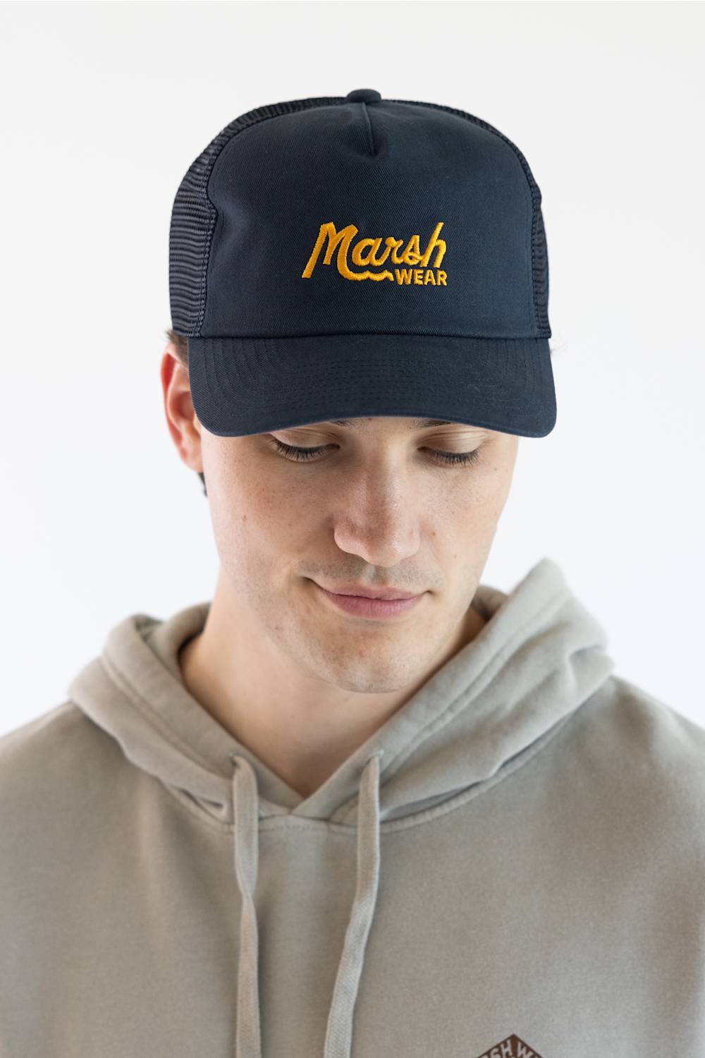 A man wearing a dark navy blue trucker hat with yellow embroidered 'Marsh Wear' text, paired with a light beige hoodie, against a plain white background.