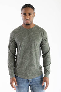 Front studio portrait of a model wearing the long-sleeve Marsh Wear Surfside Waffle Knit Shirt in Fossil Foxhole Camo green, featuring a crew neck with a cross-stitch detail and waffle-textured fabric, styled with blue jeans.