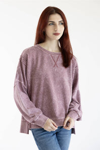 A woman with red hair wearing a dusty mauve-purple long-sleeve crewneck sweatshirt and blue jeans, standing against a plain white studio background.