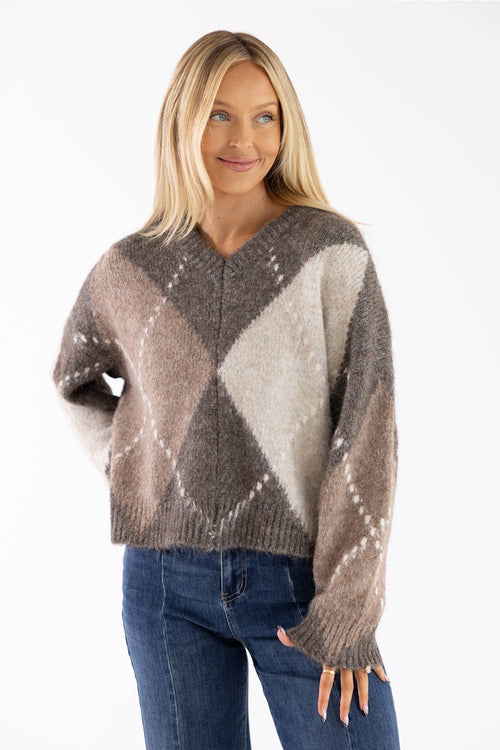 Front view of a smiling woman with long blonde hair wearing a brown Miracle argyle V-neck sweater featuring a soft fuzzy knit fabric and an all-over diamond pattern, paired with dark denim jeans.