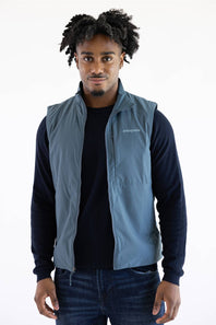 A man wearing a slate blue zip-up vest over a dark navy long-sleeve shirt and blue jeans, standing against a plain white background.