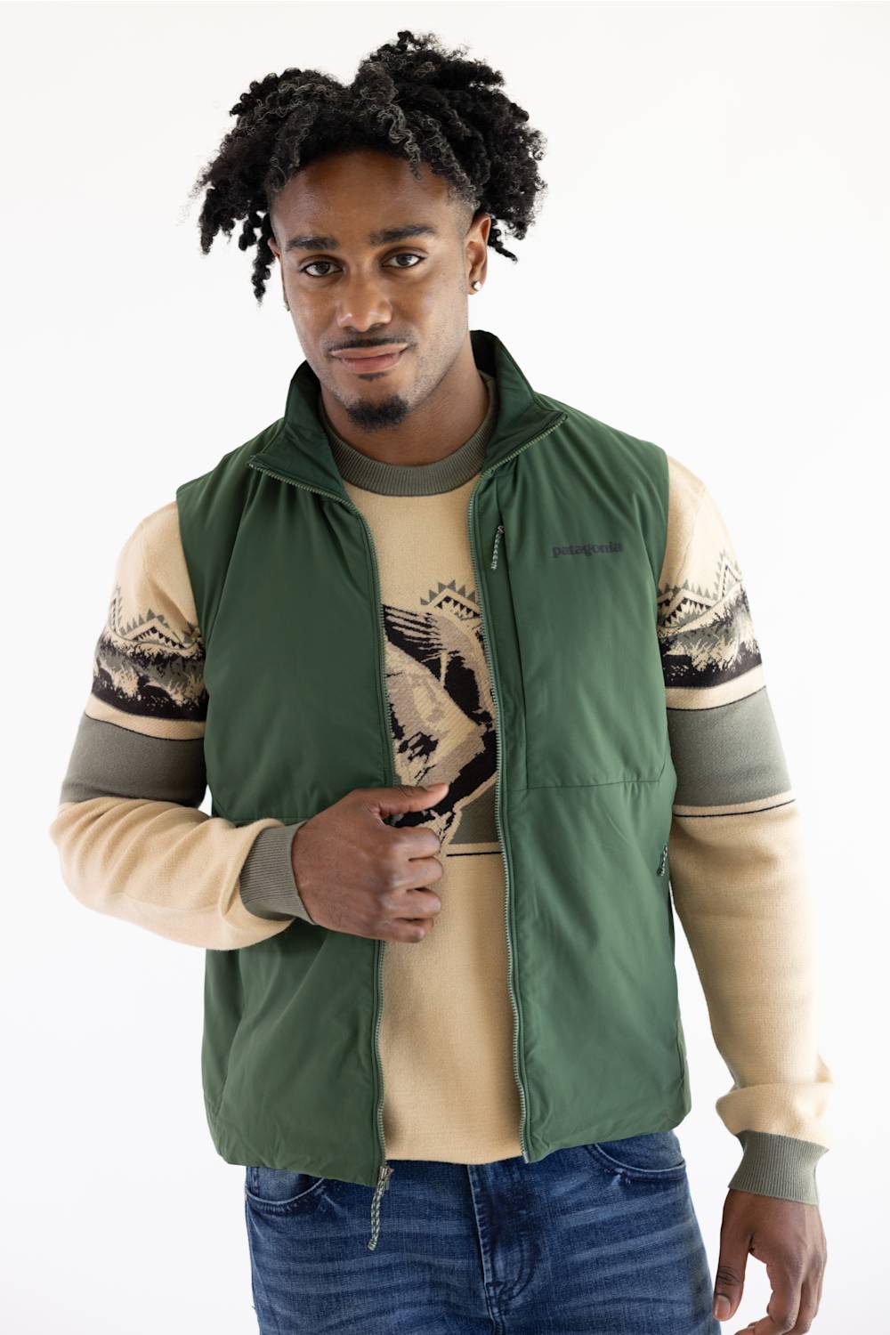 A man wearing a green zip-up vest over a beige and gray patterned long-sleeve shirt, paired with blue jeans, against a plain white background.