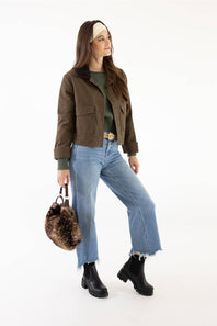 A woman modeling a brown cropped utility jacket with a dark brown faux-fur collar, styled with a green top, light-wash wide-leg jeans, black boots, and a brown furry handbag against a white studio background.