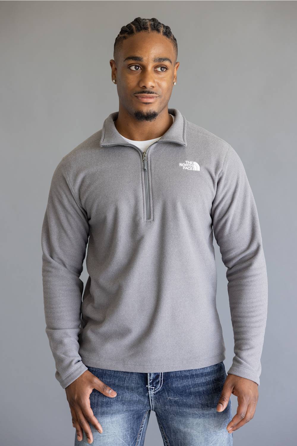 A man wearing a light gray quarter-zip fleece pullover by The North Face, paired with blue jeans, standing against a solid gray studio background.