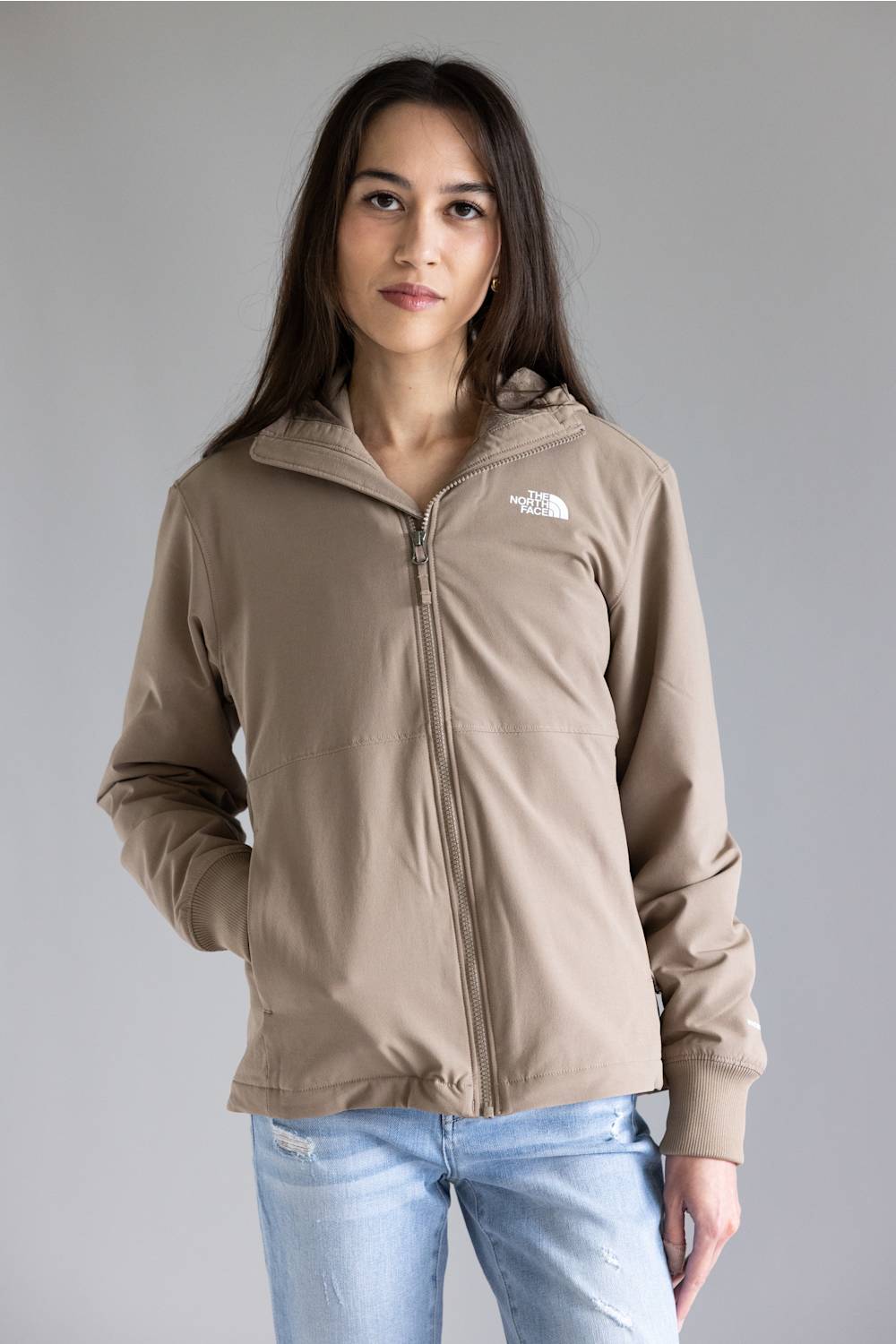 A woman wearing a light brown zip-up hoodie with a small white embroidered logo on the left chest, paired with light blue jeans, against a solid gray studio background.