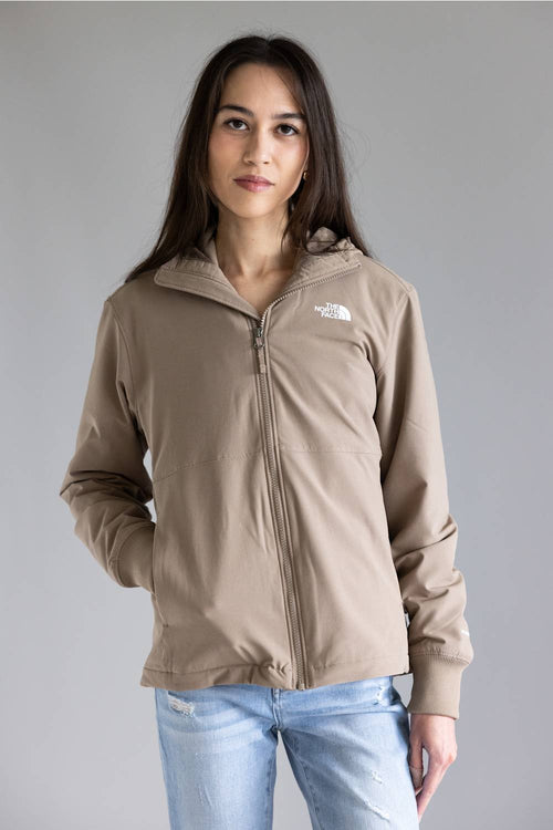 A woman wearing a light brown zip-up hoodie with a small white embroidered logo on the left chest, paired with light blue jeans, against a solid gray studio background.