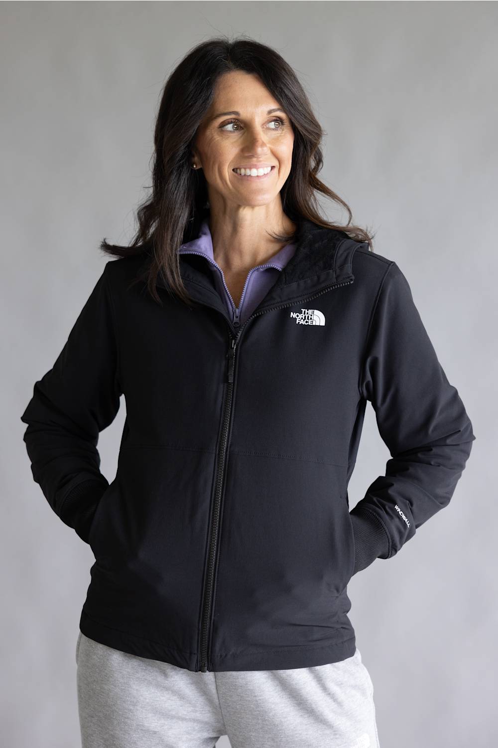 A woman wearing a black zip-up jacket by The North Face over a light purple collared shirt and light gray sweatpants, standing against a plain gray studio background.