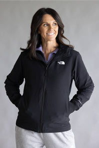 A woman wearing a black zip-up jacket by The North Face over a light purple collared shirt and light gray sweatpants, standing against a plain gray studio background.