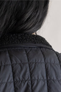 Close-up back view of a person wearing a black quilted jacket with a textured black fleece collar, showing dark hair against a light neutral background.