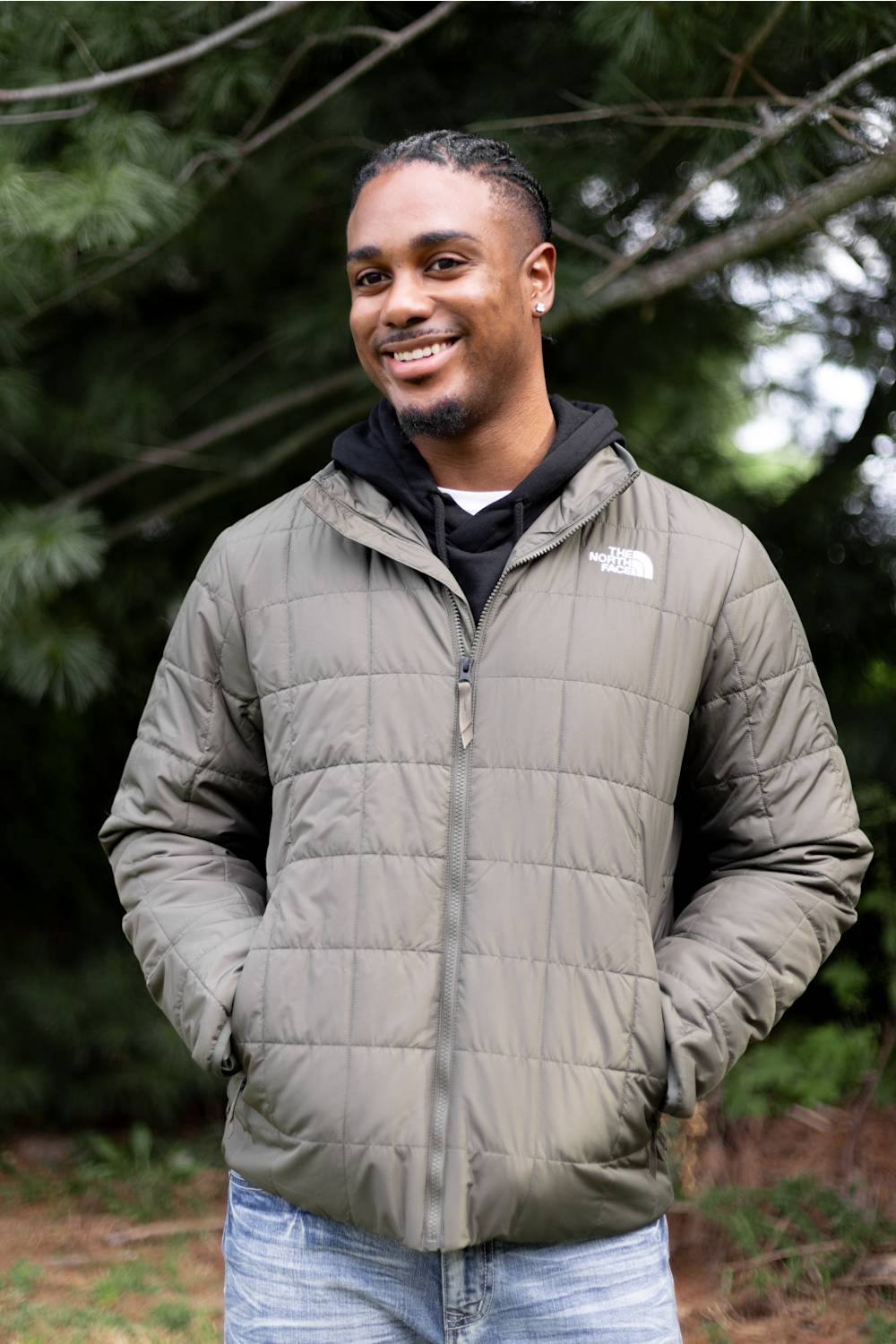A man wearing an olive green puffer jacket by The North Face, layered over a black hoodie and blue jeans, standing outdoors in front of a forest background.
