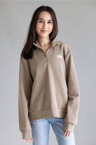 A woman wearing a taupe brown quarter-zip pullover sweatshirt by The North Face, paired with light blue jeans, standing against a neutral gray studio background.